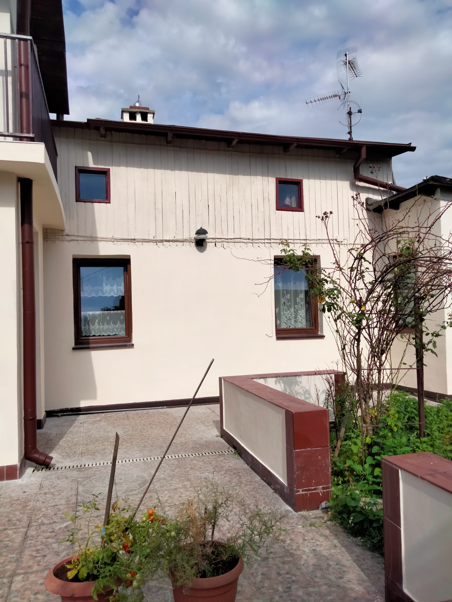 Elewacja domu jednorodzinnego po odświeżeniu, z beżowymi ścianami, brązowymi ramami okiennymi i rynnami. Widoczne elementy drewniane na górnej części fasady i antena na dachu. Na pierwszym planie...