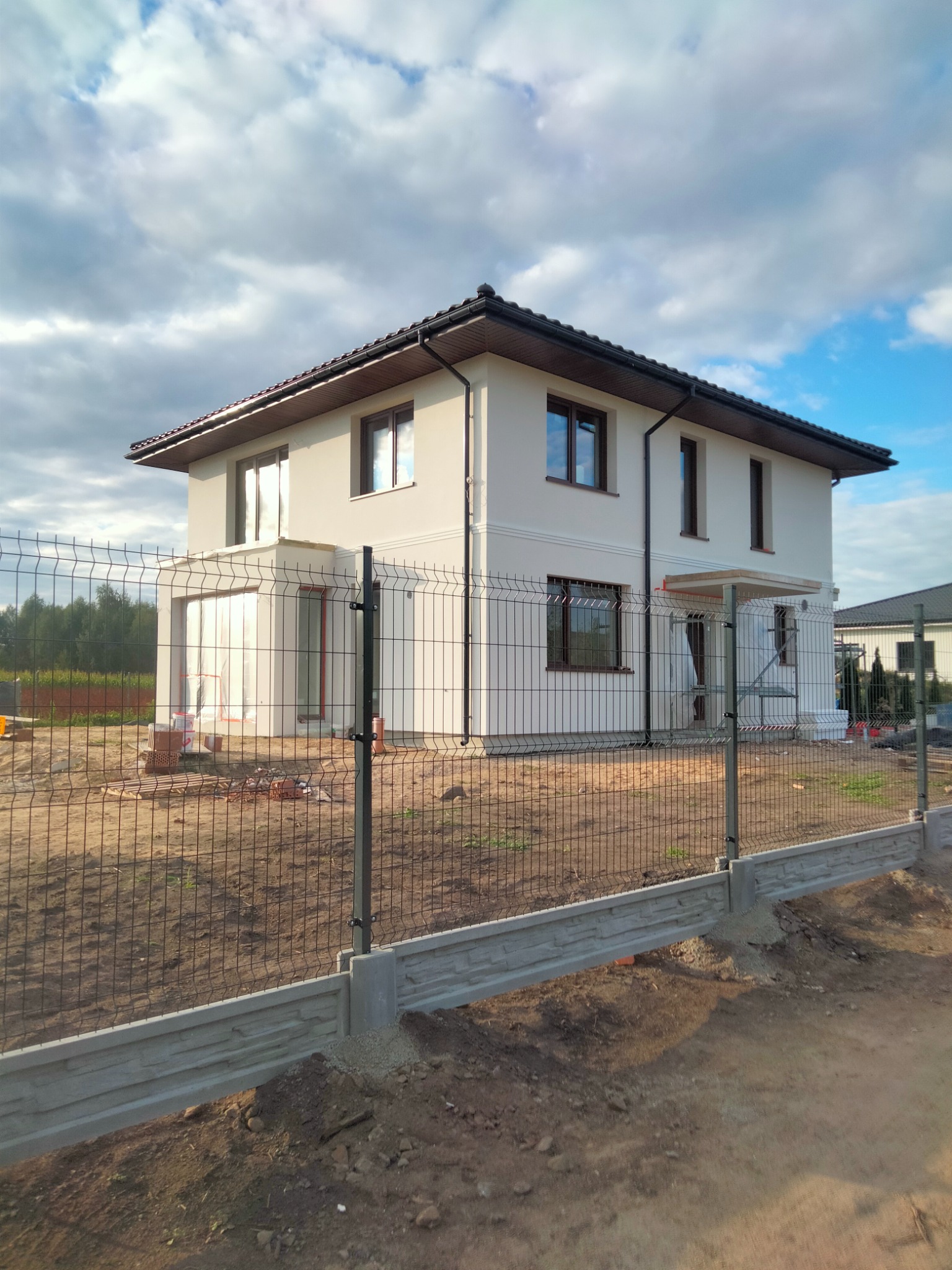 Dwukondygnacyjny dom jednorodzinny z jasną elewacją, widoczny przez metalowe ogrodzenie panelowe, w trakcie prac wykończeniowych.