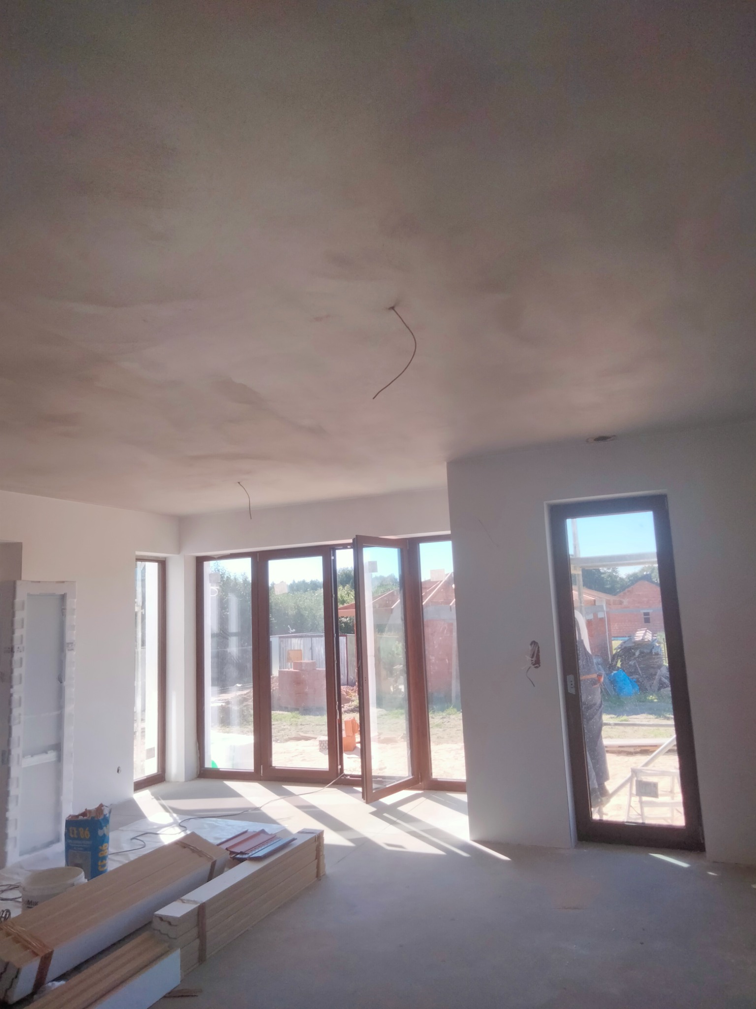 Wnętrze w trakcie prac wykończeniowych w Pabianicach: surowy sufit po szpachlowaniu, przygotowane panele podłogowe i duże okna z widokiem na zewnątrz.