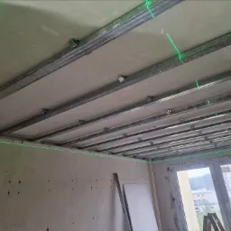 Montaż sufitu podwieszanego z płyt kartonowo-gipsowych na metalowej konstrukcji, widoczne zielone linie lasera wyznaczające poziom, surowe ściany w tle.