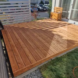 Nowoczesny taras z drewna kompozytowego, z widoczną strukturą desek, oświetlony promieniami słońca przechodzącymi przez ażurową konstrukcję, z drewnianą ławką ze skrzynią i narzędziem...