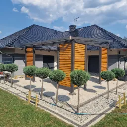 Nowoczesna altana z drewnianymi panelami i czarną konstrukcją, otoczona ozdobnymi drzewkami w kształcie kul, na tle domu z ciemnym dachem