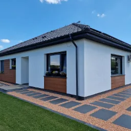 Nowoczesny dom jednorodzinny z ciemną dachówką, białymi ścianami i elementami dekoracyjnymi z paneli drewnopodobnych, otoczony trawnikiem i ścieżką z płyt na żwirze.