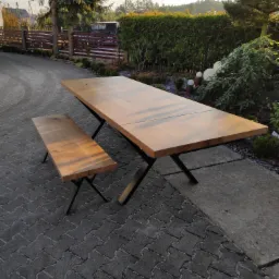 Rozkładany stół z jasnego drewna i dopasowana ławka na czarnych, metalowych nogach typu X, ustawione na kostce brukowej przed zielonym żywopłotem i ogrodzeniem.