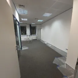 Biuro przygotowane do malowania: ściany zabezpieczone taśmą malarską, podłoga przykryta folią, widoczna drabina aluminiowa.