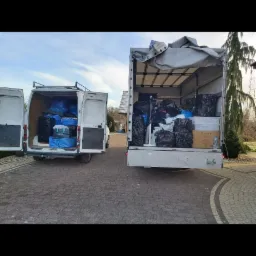 Dwa busy załadowane workami i kartonami podczas dnia. Jeden bus jest biały i w pełni załadowany, drugi bus jest biały i częściowo załadowany. Na zewnątrz widać kostkę brukową i dom w tle.