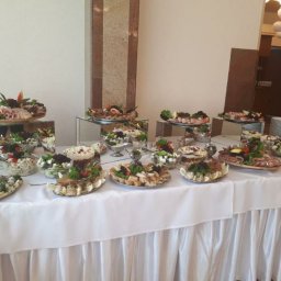 Zoja Dan sp. Z o.o - Bogato zastawiony stół cateringowy z różnorodnymi sałatkami, przekąskami mięsnymi i dekoracjami z ziół, prezentowany na lustrzanych podwyższeniach, z białym obrusem z falbanami.