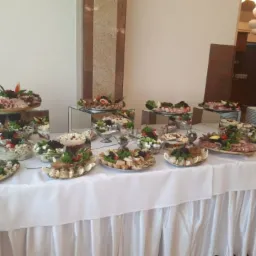Bogato zastawiony st&oacute;ł cateringowy z r&oacute;żnorodnymi sałatkami, przekąskami mięsnymi i dekoracjami z zi&oacute;ł, prezentowany na lustrzanych podwyższeniach, z białym obrusem z falbanami.