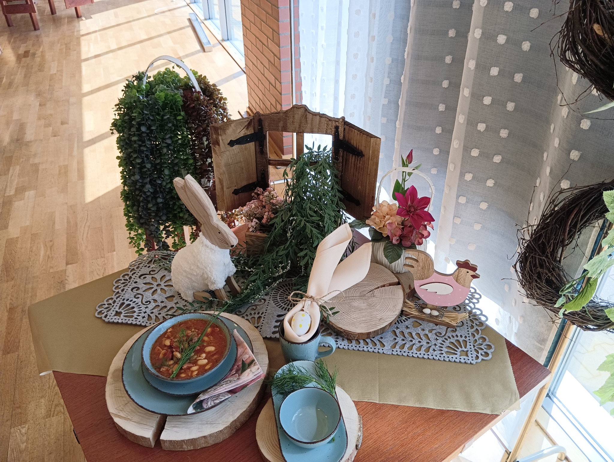 Świąteczny stół wielkanocny z zupą fasolową, dekoracjami z zajączkiem, kurczakiem, drewnianymi podkładkami i serwetkami w kształcie uszu królika, na tle drewnianej ściany i okna z firanką w kropki.