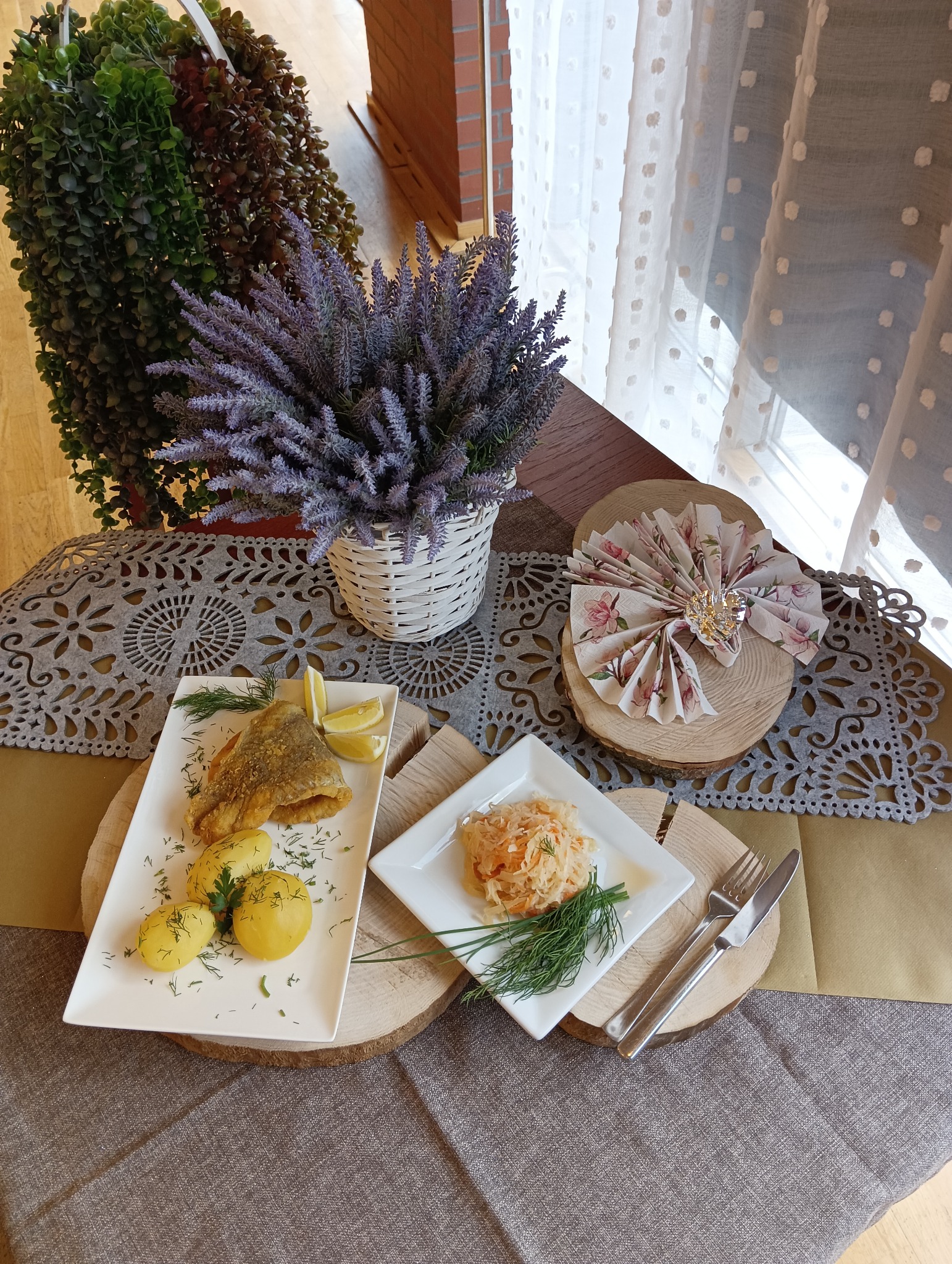 Elegancko podane danie z smażonej ryby, ziemniaków i surówki na drewnianych podkładkach, z dekoracją z lawendy i serwetką origami na tle okna.