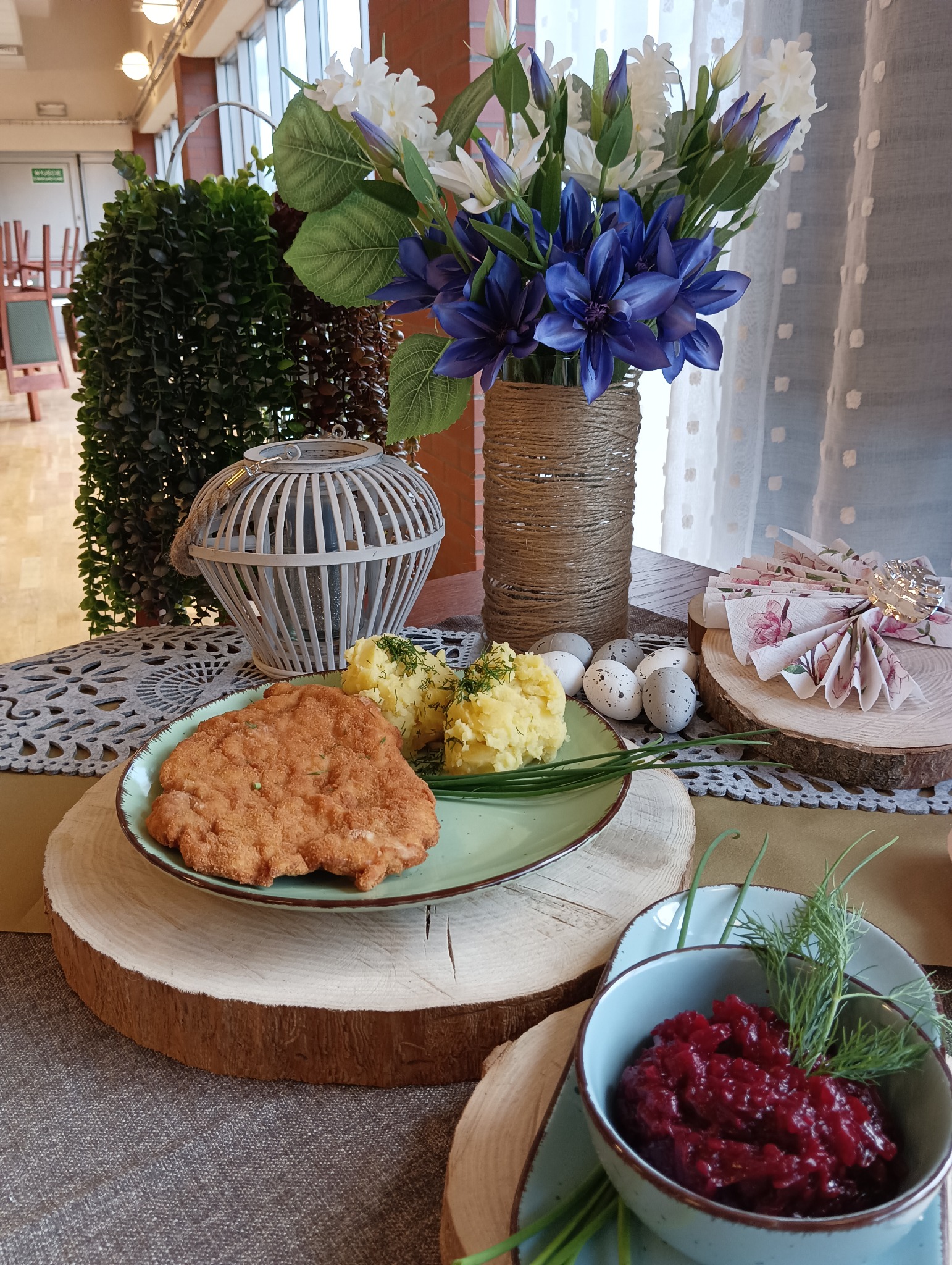 Uroczysty stół z kotletem schabowym, ziemniakami z koperkiem, ćwikłą i dekoracjami świątecznymi, w tym barwione jaja i kwiaty w jutowym wazonie.