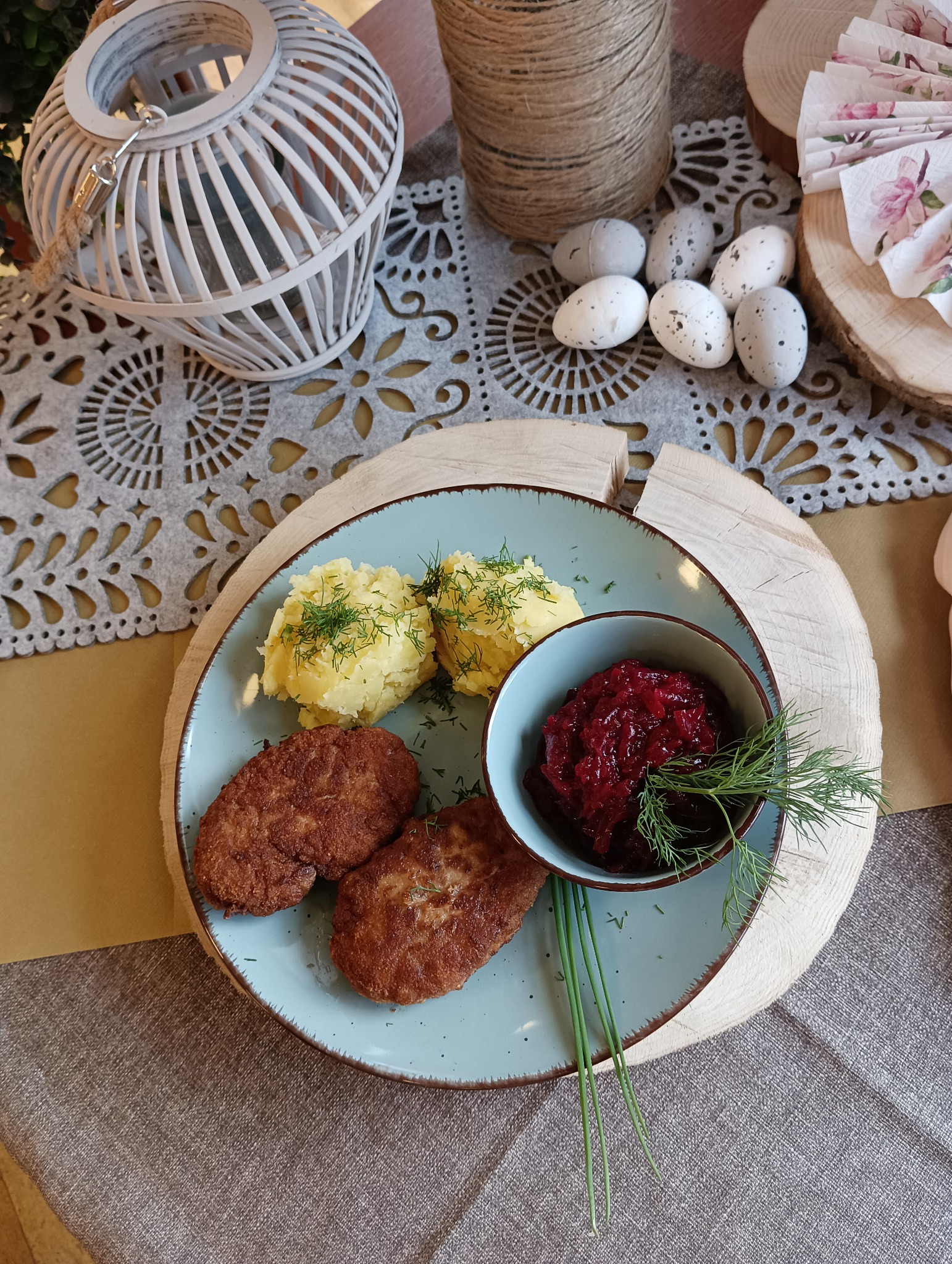 Świąteczny obiad na desce z drewna: kotlety mielone, ziemniaki z koperkiem i buraczki, dekoracja z jajek i lampionu na stole w stylu rustykalnym.