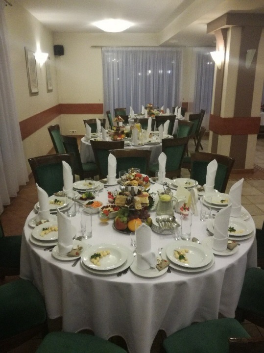 Uroczysta sala bankietowa z okrągłymi stołami nakrytymi białymi obrusami, zielonymi krzesłami i centralnie ustawionymi paterami z owocami i przekąskami, zastawą stołową i szklankami.