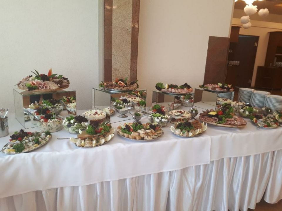 Bogato zastawiony stół cateringowy z różnorodnymi sałatkami, przekąskami mięsnymi i dekoracjami z ziół, prezentowany na lustrzanych podwyższeniach, z białym obrusem z falbanami.