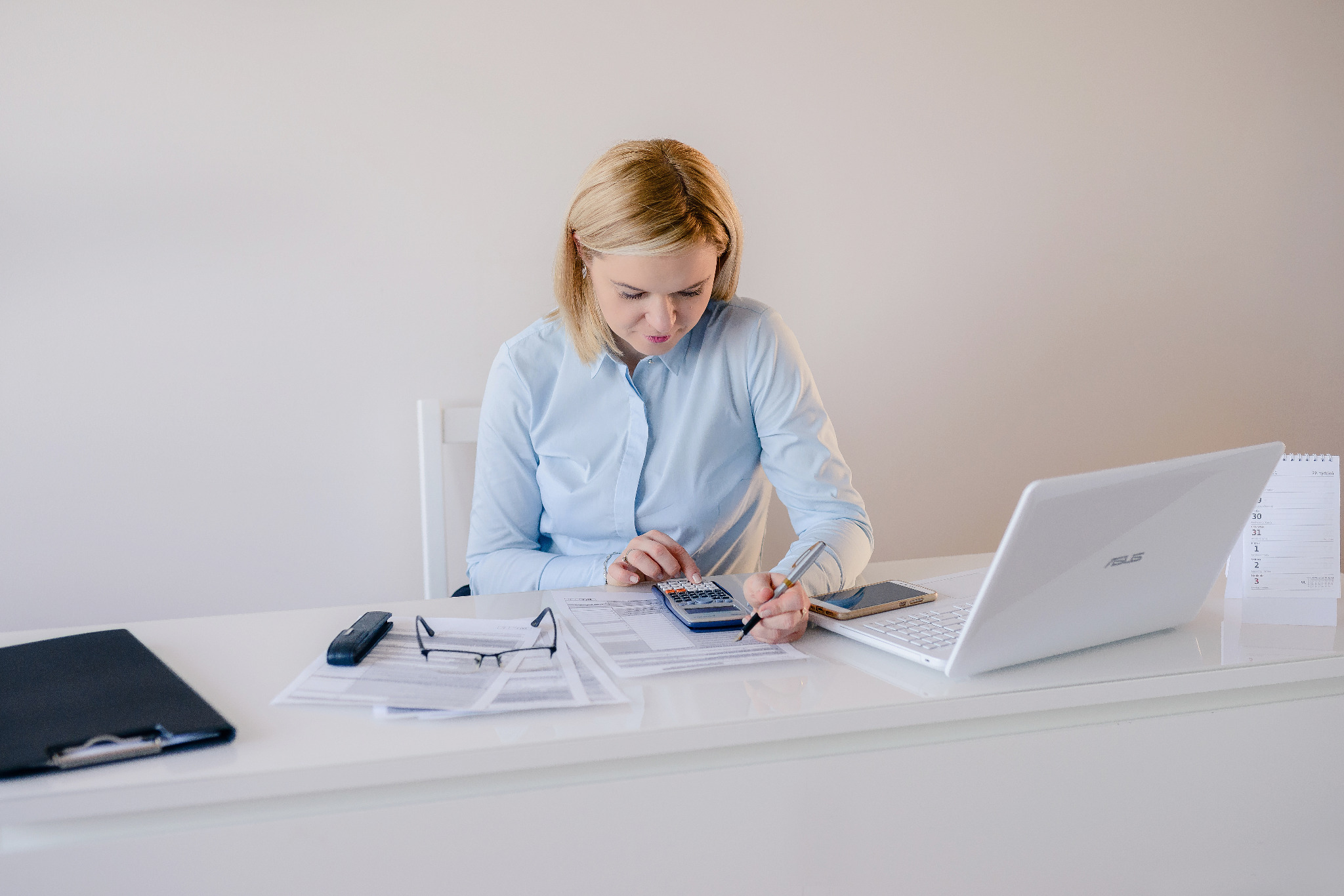 Kobieta w błękitnej koszuli przy białym biurku, obliczenia na kalkulatorze, dokumenty, laptop, telefon i kalendarz.