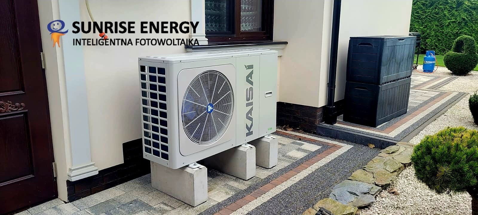 Biała jednostka zewnętrzna klimatyzacji Kaisai zamontowana na betonowych podstawach przed budynkiem z brązowymi detalami, obok widoczne czarne skrzynie ogrodowe i butla gazowa.