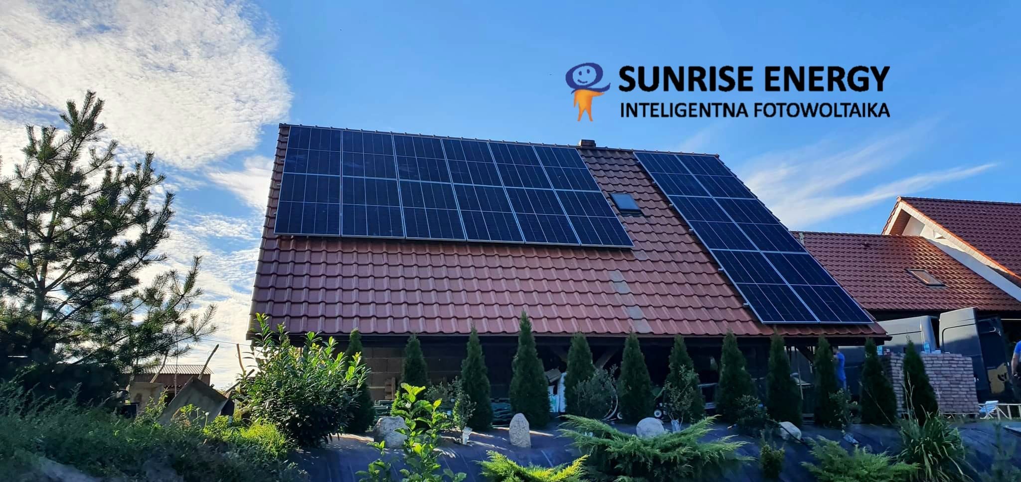 Panele fotowoltaiczne na dachu domu jednorodzinnego z czerwoną dachówką, logo firmy Sunrise Energy widoczne na górze zdjęcia.