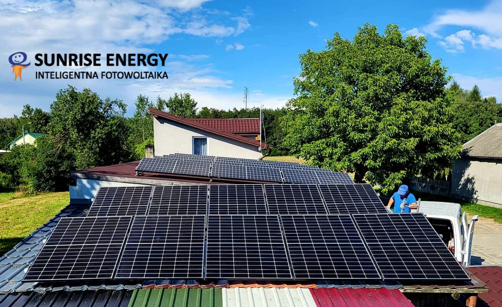 Montaż paneli słonecznych na dachu domu jednorodzinnego przez instalatora w niebieskiej koszulce, widoczny samochód dostawczy, w tle zieleń i błękitne niebo.