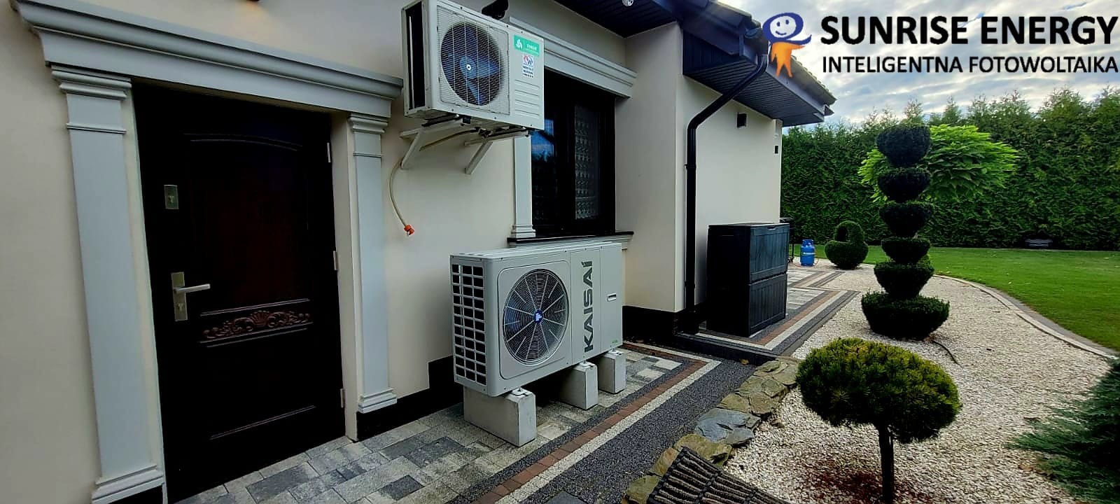 Dwa klimatyzatory Kaisai zamontowane na elewacji domu jednorodzinnego, z logo Sunrise Energy widocznym w prawym górnym rogu, na tle zadbanego ogrodu z ozdobnymi krzewami.