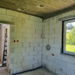 Surowy stan wnętrza budynku w trakcie prac elektrycznych: widoczne przewody elektryczne prowadzone po ścianach z bloczków, puszki instalacyjne w ścianach oraz kabel zwinięty w pętlę. Na ścianie...