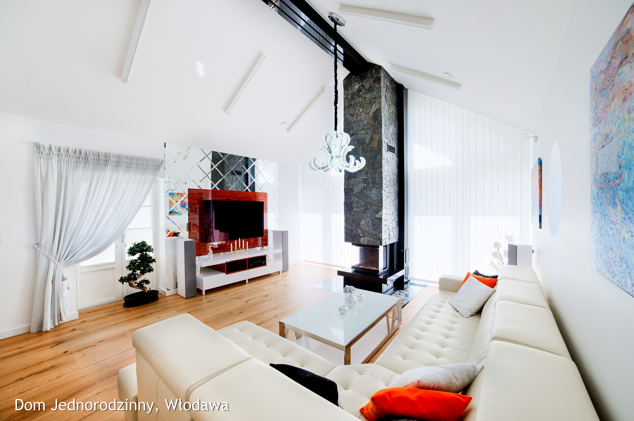 Jasny salon z drewnianą podłogą, kominkiem obłożonym kamieniem, białym narożnikiem i szklanym stolikiem kawowym, doświetlony przez okno z zasłoną i żaluzje pionowe.