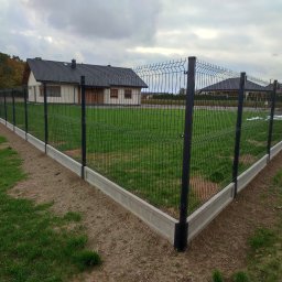 Nowoczesne ogrodzenie panelowe z podmurówką betonową, otaczające trawnik przed domem. Ciemna kolorystyka paneli kontrastuje z zielenią trawy i szarością podmurówki.