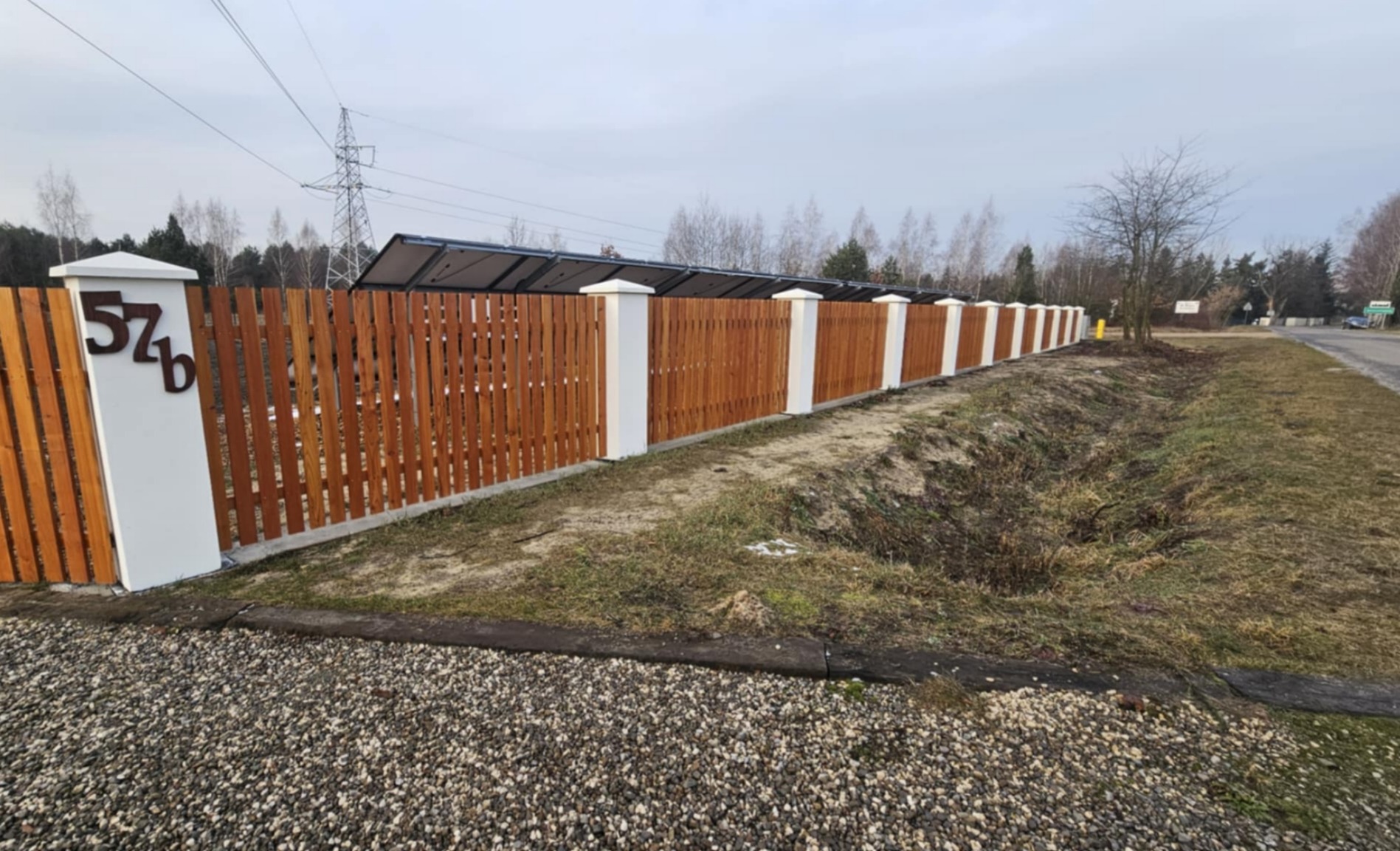 Nowoczesne ogrodzenie z drewnianymi sztachetami i białymi słupkami, z numerem domu '57b' zamontowanym na jednym ze słupków.