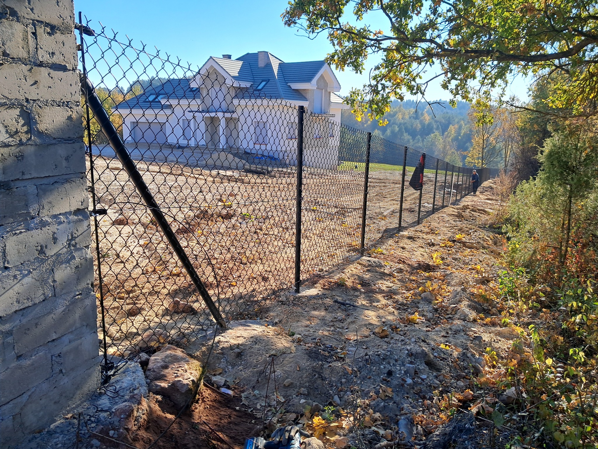 Siatka ogrodzeniowa z czarnymi słupkami na tle domu i drzew. Widoczny fragment murowanego słupka. Jesienne liście na ziemi wzdłuż ogrodzenia.