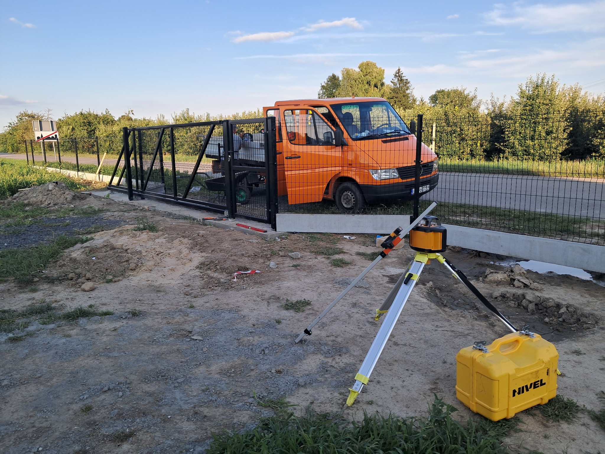 Ciemna brama przesuwna z paneli, częściowo zamontowana. Na pierwszym planie niwelator laserowy i pomarańczowy samochód dostawczy. Tło: zieleń i błękitne niebo.