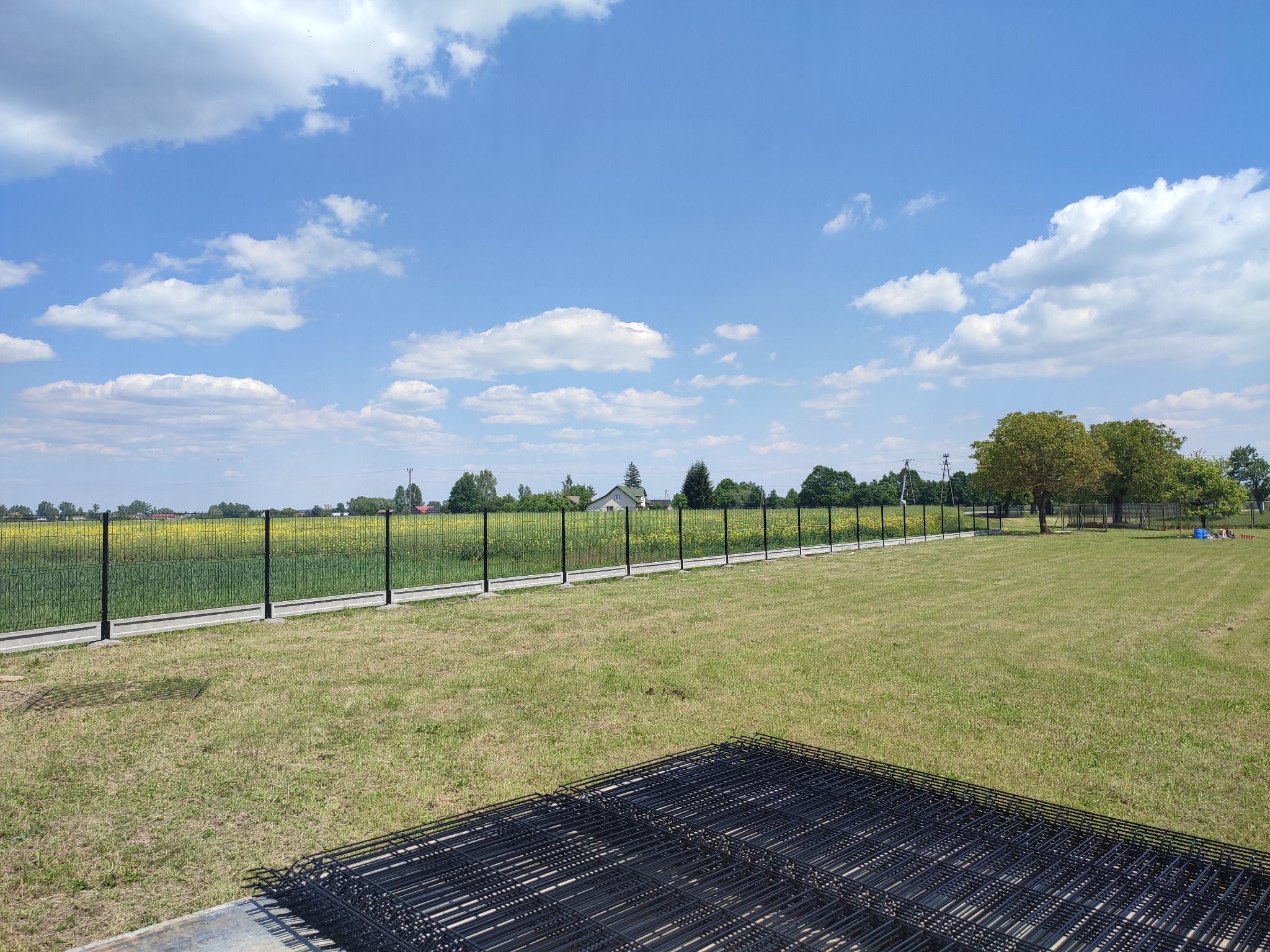 Nowoczesne, czarne ogrodzenie panelowe na betonowej podmurówce, otoczone zielenią i błękitnym niebem. Na pierwszym planie stos paneli ogrodzeniowych.