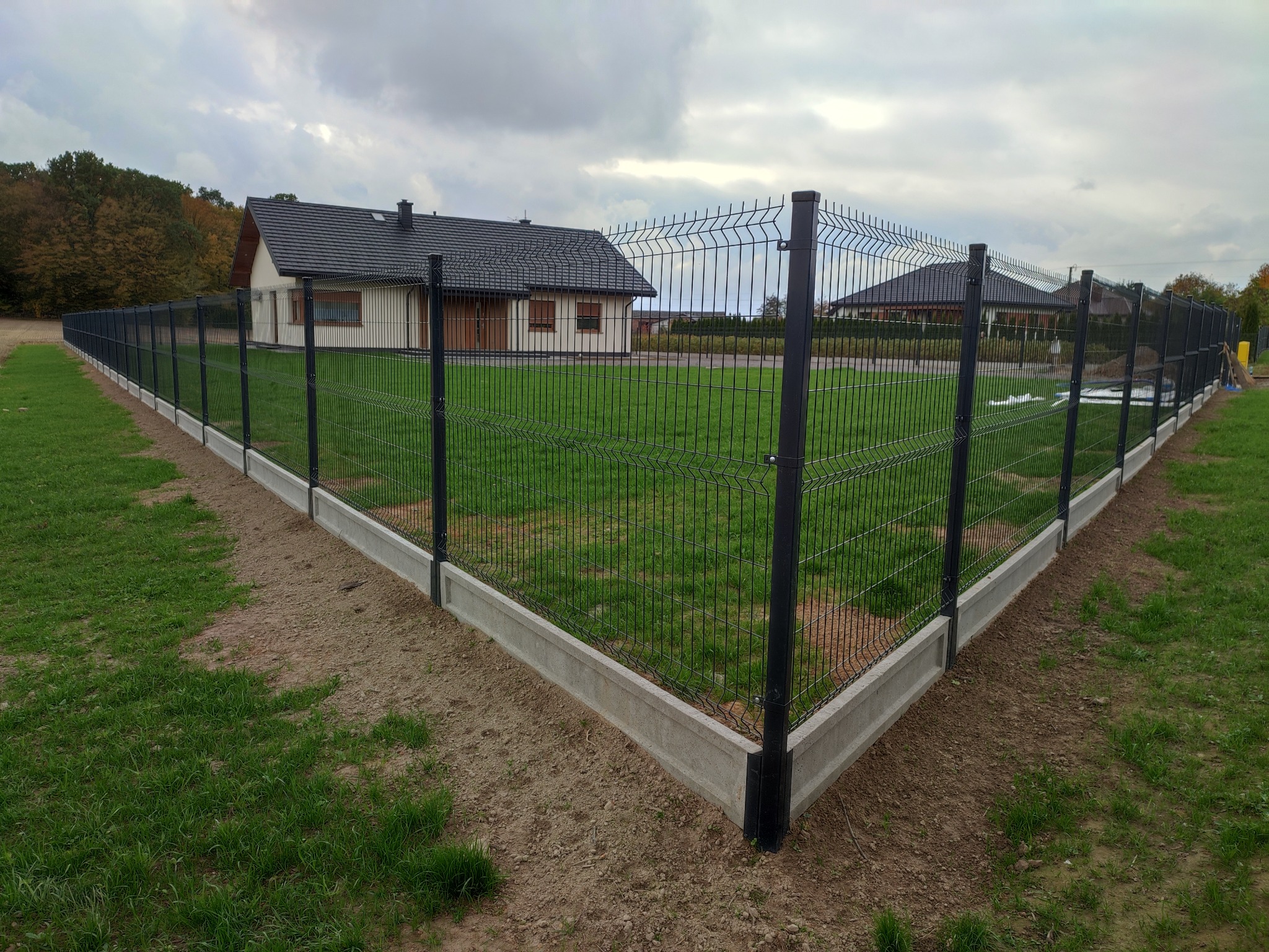 Nowoczesne ogrodzenie panelowe z podmurówką betonową, otaczające trawnik przed domem. Ciemna kolorystyka paneli kontrastuje z zielenią trawy i szarością podmurówki.