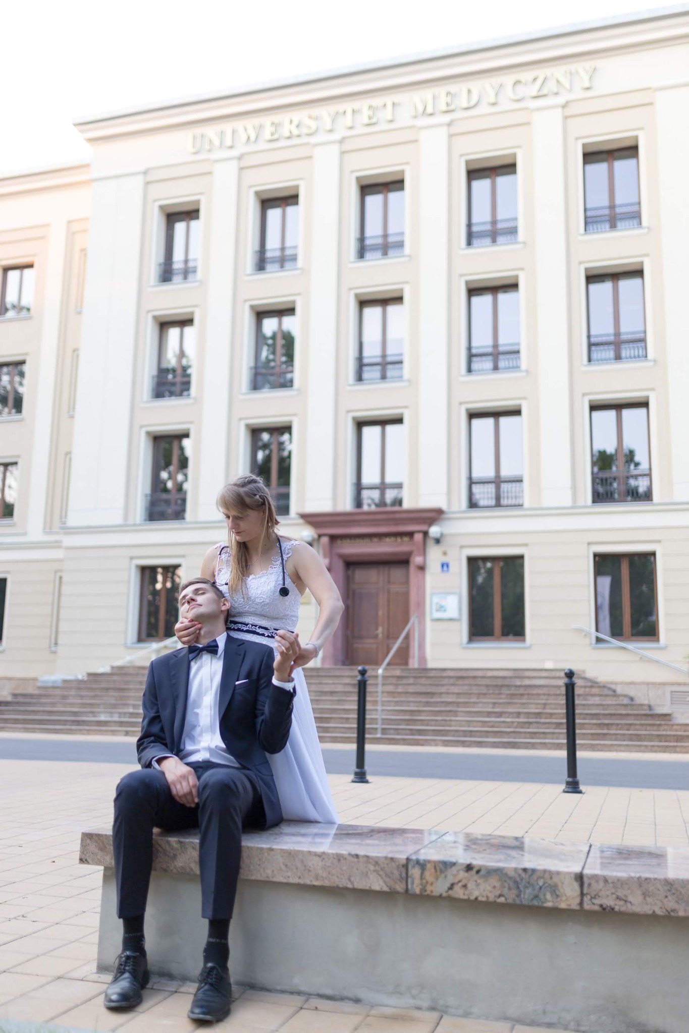 Panna młoda w białej sukni i ze stetoskopem obejmuje szyję pana młodego siedzącego na murku przed budynkiem Uniwersytetu Medycznego.