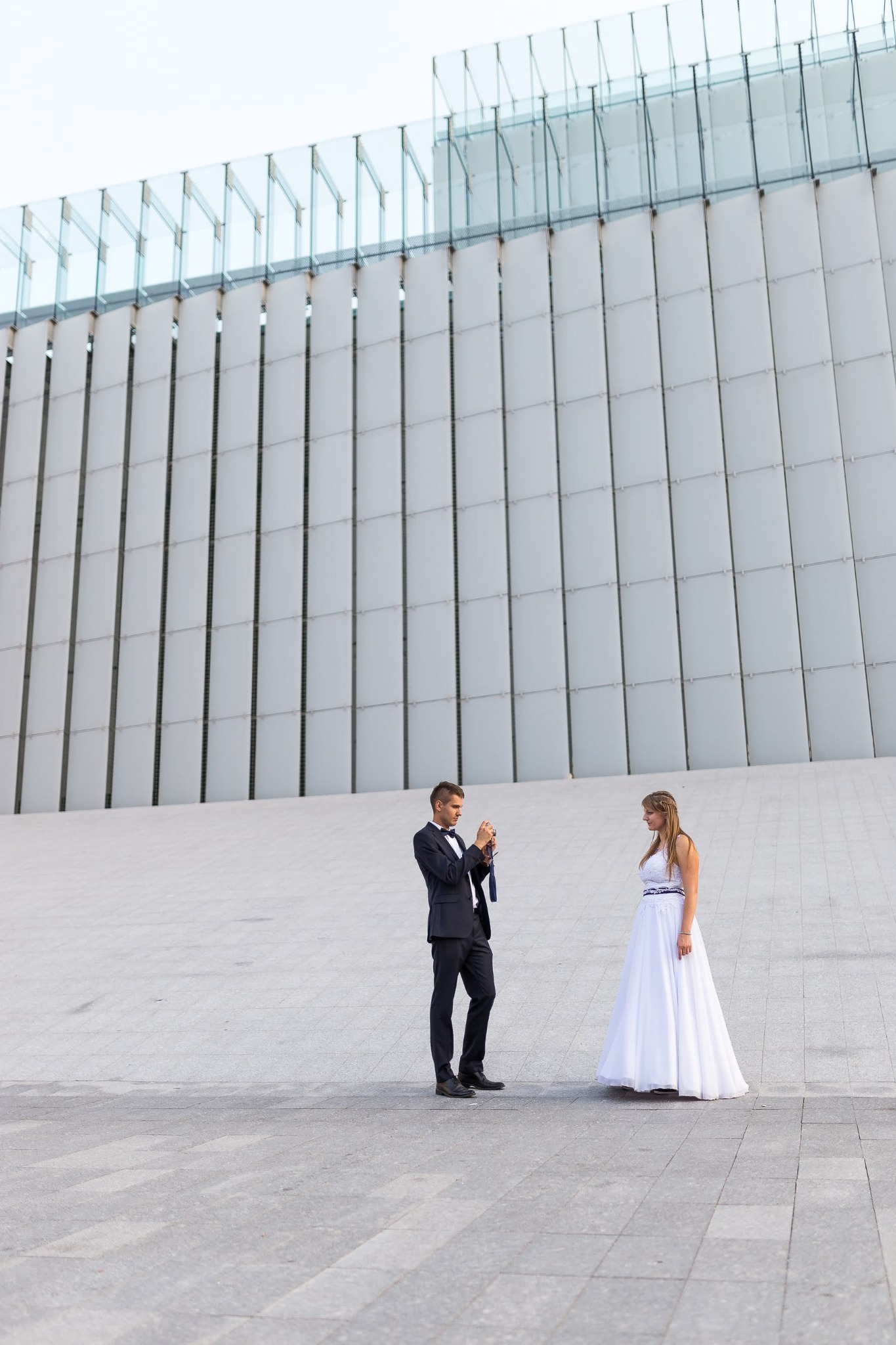 Para młoda w nowoczesnej architekturze: pan młody robi zdjęcie pannie młodej telefonem, na tle budynku z szarymi panelami i szklaną konstrukcją na górze.