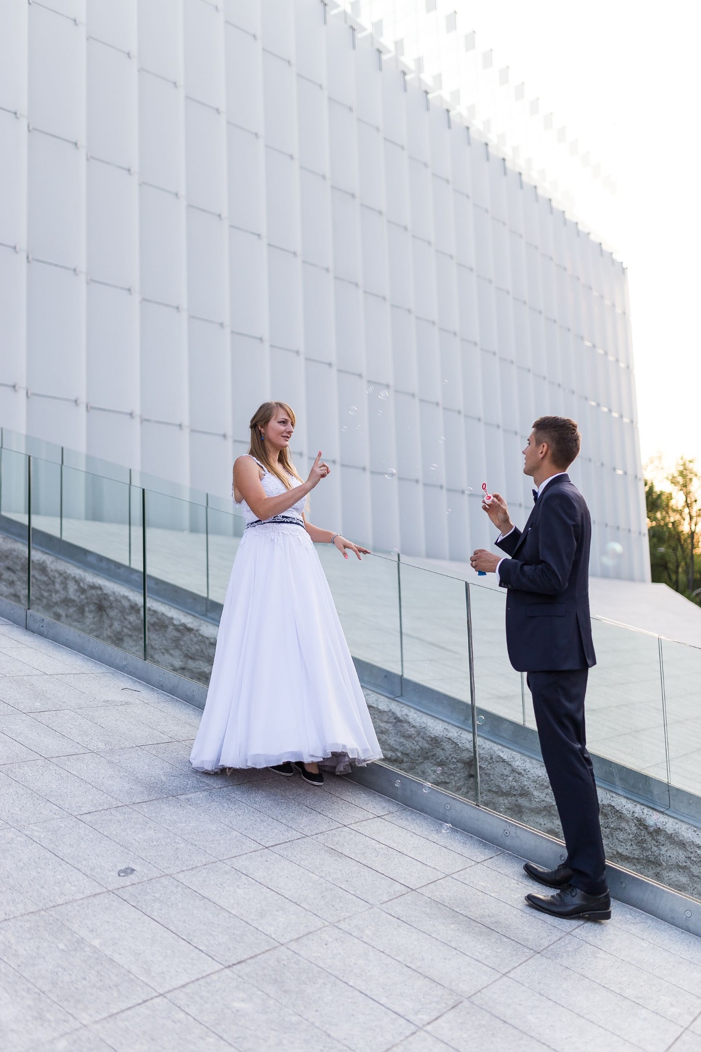Para młoda w strojach ślubnych, puszczająca bańki mydlane na tle nowoczesnej, białej architektury.