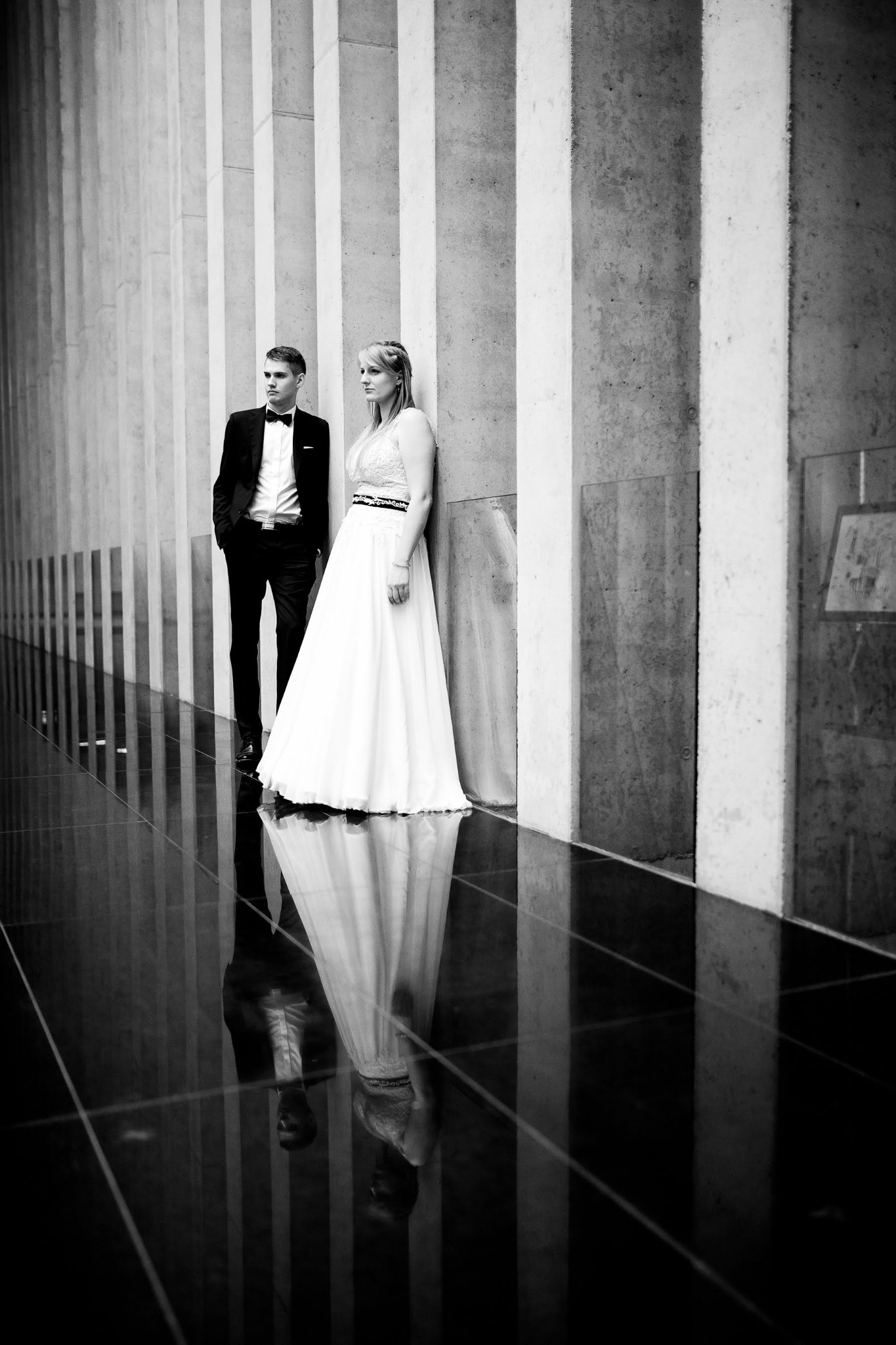 Czarno-biała fotografia pary: mężczyzna w smokingu i kobieta w białej sukni, opierająca się o betonową ścianę z kolumnami, z odbiciem w błyszczącej podłodze.