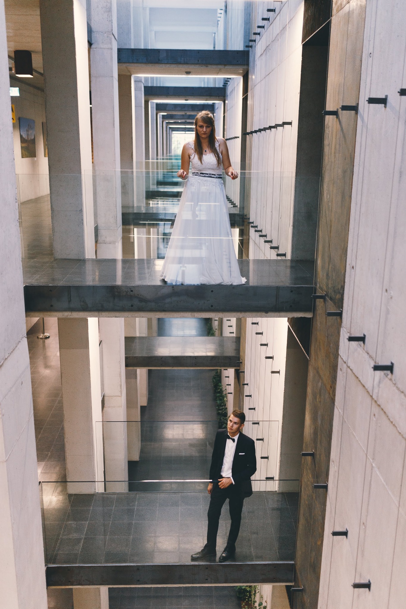 Para młoda: panna młoda w białej sukni na górnej platformie, pan młody w garniturze z muszką na dolnej, w nowoczesnym wnętrzu z betonowymi kolumnami i przeszklonymi balustradami.