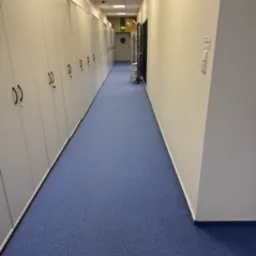 Długi korytarz wyłożony niebieską wykładziną dywanową, po lewej stronie rząd białych szaf z zamkami, na końcu korytarza drzwi z zielonym światłem ewakuacyjnym.