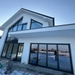 Nowoczesny dom z dużymi, lustrzanymi oknami po umyciu, widoczny w zimowym krajobrazie z resztkami śniegu na ziemi. W tle błękitne niebo. Na pierwszym planie wózek z akcesoriami do czyszczenia okien.