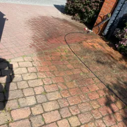 Częściowo umyta kostka brukowa z widoczną różnicą w kolorze, zarośnięta drobnymi chwastami, z wężem ciśnieniowym i fragmentem myjki przy murze z czerwonej cegły.