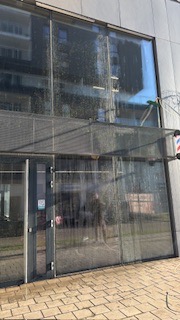 Czyszczenie szklanej fasady budynku z użyciem lancy teleskopowej. Widoczne ślady spływającej wody i piany, odzwierciedlenie otoczenia w czystej części szyby.