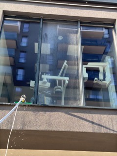 Okno z zewnątrz, częściowo umyte, z widocznym sprzętem do mycia na wysokości, odbijającym się w oknie budynku po drugiej stronie ulicy. Wewnątrz pomieszczenia widoczny sprzęt medyczny.