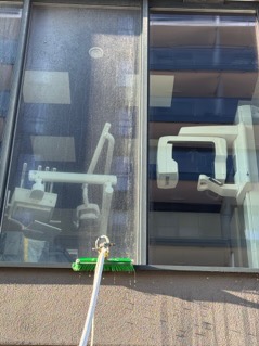 Zewnętrzna ściana budynku z dużymi oknami, na których widoczne są smugi po myciu. Na pierwszym planie zielona szczotka na długim kiju, używana do czyszczenia. Przez okna widoczny sprzęt medyczny.