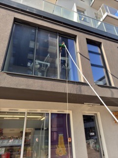 Pracownik w Łodzi myje okno na wysokości na tle szarej elewacji budynku, używając długiego kija teleskopowego z zamocowaną myjką.