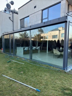 Szklana zabudowa tarasu z widocznym odbiciem otoczenia w czystych szybach, na trawniku leży rura i narzędzia, prawdopodobnie po zakończeniu prac.