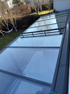 Szklany dach tarasu po umyciu, widoczny z góry, z odblaskami nieba i drzew. Na drugim planie trawnik, krzewy i biała ściana budynku.