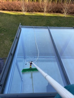 Czyszczenie szklanego dachu ogrodu zimowego z użyciem szczotki teleskopowej, widok z góry na mokrą powierzchnię i ślady wody.