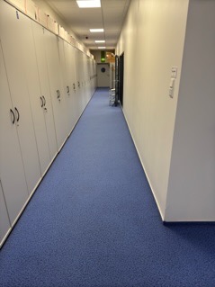 Długi korytarz wyłożony niebieską wykładziną dywanową, po lewej stronie rząd białych szaf z zamkami, na końcu korytarza drzwi z zielonym światłem ewakuacyjnym.