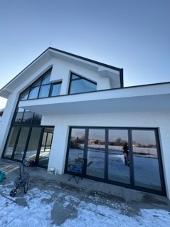 Nowoczesny dom z dużymi, lustrzanymi oknami po umyciu, widoczny w zimowym krajobrazie z resztkami śniegu na ziemi. W tle błękitne niebo. Na pierwszym planie wózek z akcesoriami do czyszczenia okien.