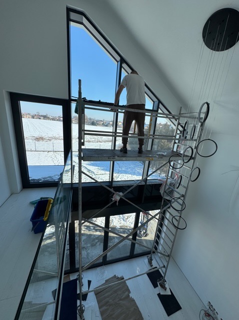 Pracownik na rusztowaniu czyści wysokie okno w nowoczesnym domu z widokiem na zaśnieżone pole; widoczny sprzęt do mycia i designerska lampa wisząca.