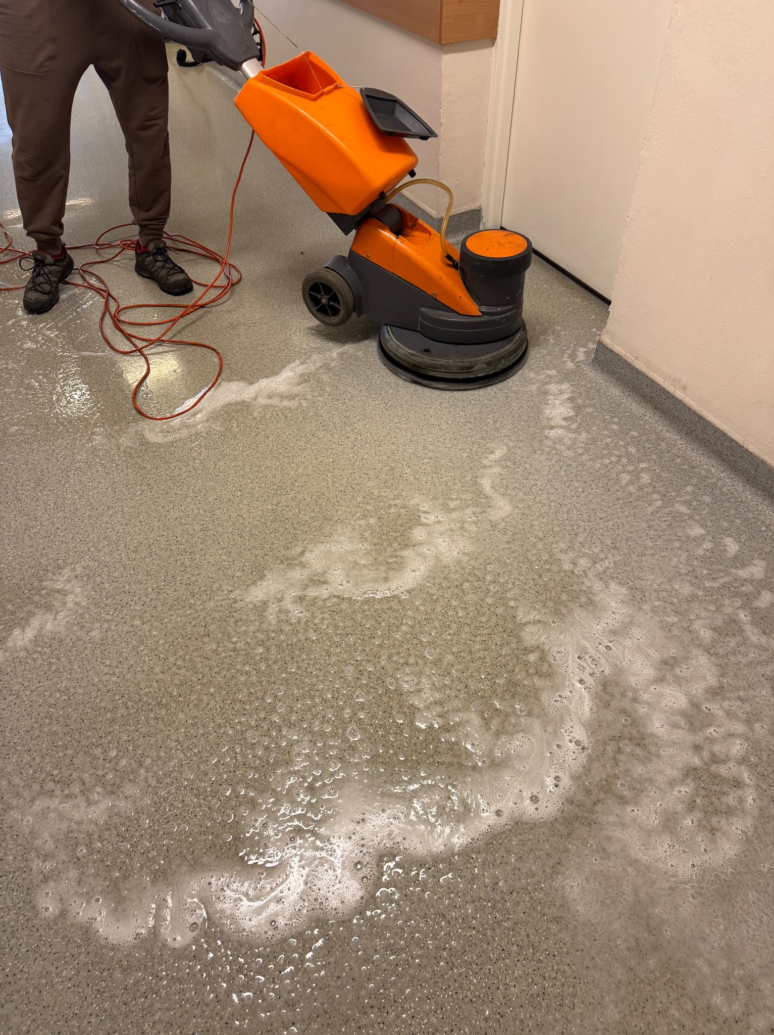 Doczyszczanie posadzki za pomocą pomarańczowej maszyny polimeryzującej, widoczne ślady detergentu i operator w brązowych spodniach.